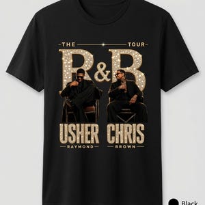 May include: A black short-sleeve t-shirt featuring a graphic print with the text THE R&B TOUR. The design includes images of two men in suits sitting in chairs, with the names USHER RAYMOND and CHRIS BROWN printed in gold glitter-style lettering.