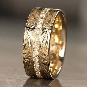 May include: A wide gold band ring featuring a central vertical row of small, round, clear gemstones. The surface of the gold band is decorated with intricate, engraved scrollwork patterns on both sides of the central stone setting.