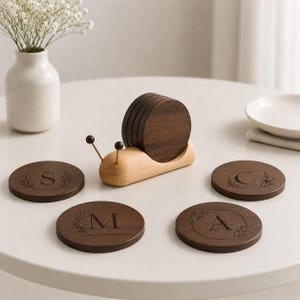 May include: A set of circular dark brown wooden coasters featuring engraved letters S, M, C, and A, each surrounded by a delicate floral wreath design. The coasters are stored in a light-colored wooden snail-shaped holder with two thin antennae.