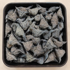 Puede incluir: Una colección de frutos de castaña de agua secos, de color gris oscuro, con protuberancias afiladas en forma de cuerno, dispuestos en un recipiente cuadrado de color marrón oscuro. Cada fruto tiene una textura leñosa y forma irregular.
