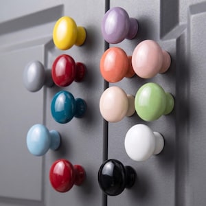 May include: A collection of twelve round, glossy cabinet knobs in various solid colors including yellow, red, blue, light blue, purple, orange, pink, cream, light green, white, black, and gray, mounted on a dark gray paneled cabinet door surface.