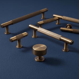 May include: A collection of antique brass cabinet hardware including several long bar pulls, a T-shaped handle, and a single round knob. Each piece features a textured, ribbed center section with smooth, cylindrical ends and circular mounting bases.
