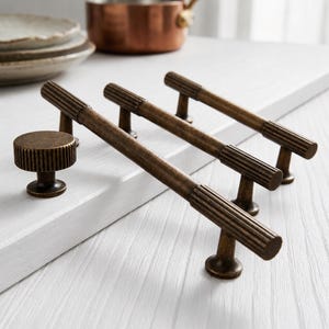 Könnte beinhalten: Ein Set aus antikem Messingbeschlag, bestehend aus einem runden Knopf und drei länglichen Schubladengriffen. Jedes Teil weist ein texturiertes, geripptes Design an den Kanten und ein dunkles, verwittertes Metallfinish für Schränke auf.