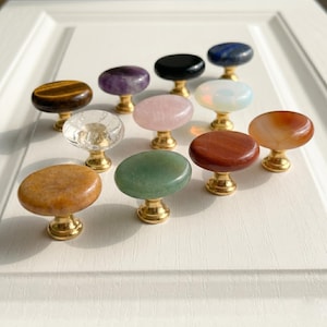 Puede incluir: Una colección de once pomos para armarios con cabezas de piedra natural lisa de forma ovalada en varios colores, incluyendo marrón, morado, negro, azul, rosa, blanco, naranja, verde y transparente, montados sobre una base de metal dorado.