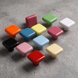 Könnte beinhalten: Eine Sammlung von zwölf quadratischen Schrankknöpfen in verschiedenen Unifarben, darunter Rot, Orange, Gelb, Grün, Blau, Rosa, Schwarz und Weiß. Jeder Knopf hat eine glatte, glänzende Oberfläche mit abgerundeten Ecken und einem zentralen Sockel.