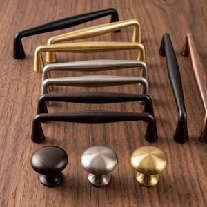 Könnte beinhalten: Eine Sammlung von Möbelbeschlägen, bestehend aus mehreren gewölbten Metall-Schubladengriffen in Mattschwarz, gebürstetem Gold, Silber und Bronze sowie drei runden Schrankknöpfen in Dunkelbraun, Silber und gebürstetem Gold.