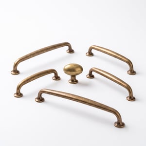 May include: A collection of five antique brass-finished cabinet hardware pieces, including four arched drawer pulls of varying lengths and one central oval-shaped cabinet knob, all featuring a smooth, metallic surface with subtle dark patina.