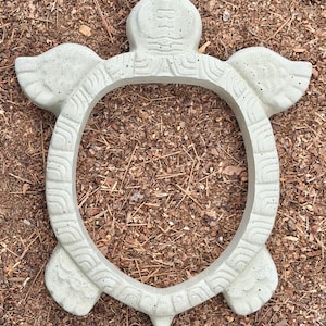 May include: A light gray concrete turtle-shaped garden ring with a hollow center. The outer rim features a repeating curved pattern, while the head and four flippers are textured with subtle, molded details. The surface has a smooth, matte finish.