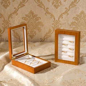 May include: Two wooden ring display boxes with glass lids and white velvet inserts. One box is open, revealing five gold rings in slots, while the other is closed and upright, displaying five gold rings arranged in horizontal rows within the case.