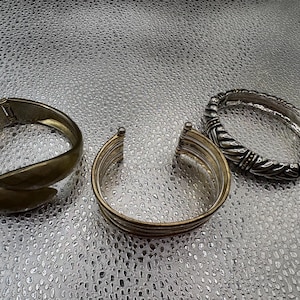 Puede incluir: Tres pulseras se muestran. Una es un brazalete articulado de tono dorado mate y liso. La segunda es un brazalete abierto con múltiples bandas finas superpuestas en tonos dorados y plateados. La tercera es una pulsera articulada de tono plateado oxidado.