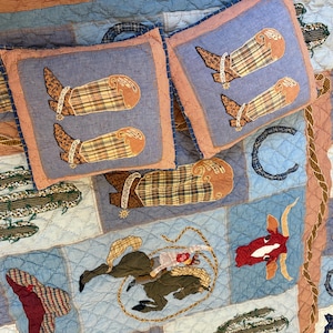 May include: A quilted bedding set featuring two square pillows and a larger quilt decorated with western-themed patchwork patterns including cowboy boots, cacti, horses, longhorn skulls, horseshoes, and stars in shades of blue, brown, and tan.