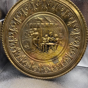 May include: A round brass decorative plate featuring an embossed scene of four figures gathered around a table in an interior setting. The border is decorated with a repeating scroll pattern, and the central relief depicts a fireplace and furniture.
