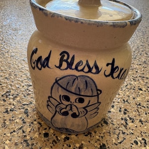 May include: A beige ceramic canister with a matching lid, featuring a blue hand-painted illustration of an angel with a halo and the text God Bless Texas in blue script. The rim and base have a speckled blue decorative trim finish.