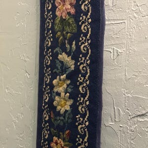 May include: A long, vertical needlepoint wall hanging featuring a deep navy blue background. The centre displays a repeating floral pattern with pink, yellow, and white flowers and green leaves, framed by intricate cream-coloured scrollwork borders.