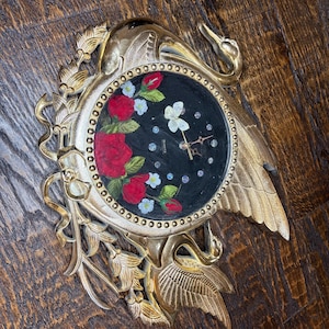 May include: A gold-toned wall clock featuring two swan figures with ornate wing details. The circular clock face is black with red rose illustrations, small white flowers, a white butterfly, and clear crystal hour markers. The word QUARTZ is printed.