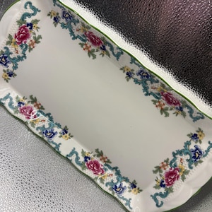 May include: A rectangular white ceramic serving tray with scalloped edges and a green rim. The border features a repeating pattern of pink peonies, small blue and yellow flowers, and intricate teal scrollwork accents on a white background.