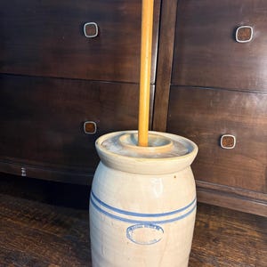 Puede incluir: Una mantequera cilíndrica de gres beige con dos rayas azules horizontales y un sello ovalado azul que dice Marshall Pottery Texas. Incluye una tapa ajustada con un orificio central y un mango de madera largo y liso para batir.