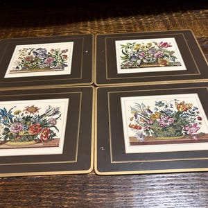May include: A set of four rectangular placemats featuring dark brown borders with thin gold trim. Each placemat displays a central rectangular illustration of a colorful floral arrangement in a basket, set against a cream-colored background.