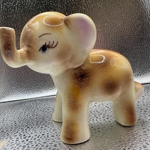 May include: A ceramic elephant figurine featuring a glossy cream-colored base with brown airbrushed spots across the body. The elephant has a raised trunk, painted blue eyes with long black eyelashes, and large ears with a subtle pinkish tint.