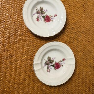 Può includere: Due posacenere rotondi in porcellana bianca con un delicato motivo floreale di rose rosa e foglie verdi al centro. Ogni posacenere presenta un bordo smerlato e testurizzato con un sottile profilo metallico dorato e due incavi per appoggio.