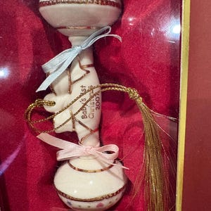 May include: A white ceramic baby rattle ornament with gold trim, featuring pastel-colored hearts and stars. The rattle is inscribed with the text Baby's First Christmas 2007 and is decorated with a light blue ribbon, a pink ribbon, and a gold tassel.