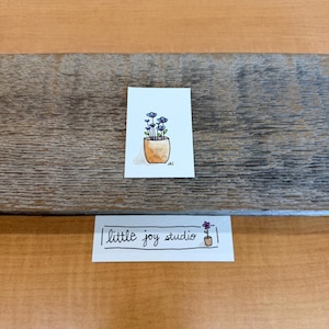 May include: A small rectangular card featuring a watercolor illustration of blue flowers in a tan pot, accompanied by a separate white rectangular label with the black handwritten text little joy studio and a small drawing of a flower in a pot.