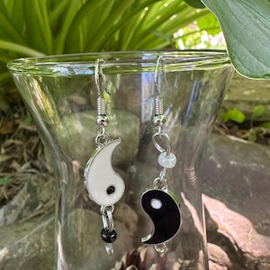 Puede incluir: Un par de pendientes colgantes con símbolos de yin y yang. Un pendiente tiene un dije de yin yang blanco con un punto negro y una pequeña cuenta negra. El otro pendiente tiene un dije de yin yang negro con un punto blanco y una pequeña cuenta transparente.