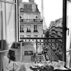 May include: A black and white scene showing a person in a white bathrobe reading a Le Figaro newspaper. A breakfast tray with croissants, bread, and white ceramic teapots sits nearby. Through the open balcony doors, Parisian buildings and the Eiffel Tower are visible.