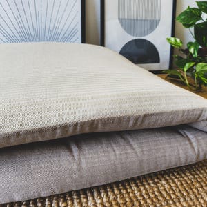 Può includere: Un cuscino da pavimento rettangolare piegato in tessuto beige chiaro con un sottile motivo a spina di pesce ripetuto. Il cuscino è spesso e morbido, mostrando un bordo stratificato distinto dove la sezione superiore sovrappone la base.