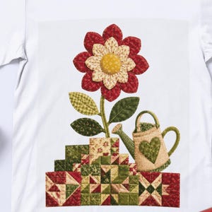 Puede incluir: Una camiseta blanca con un gráfico estilo acolchado de una flor grande con pétalos rojos y crema y un centro amarillo. Debajo de la flor hay una base de patchwork en tonos rojo, verde y crema, junto a una pequeña regadera con un corazón verde.
