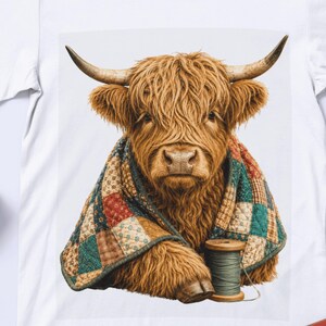 Może przedstawiać: Biały t-shirt z nadrukiem graficznym przedstawiającym kudłate brązowe szkockie bydło górskie z długimi rogami. Zwierzę jest owinięte w kolorową narzutę patchworkową w różne kwiatowe i geometryczne wzory, obok drewnianej szpulki z zieloną nicią.