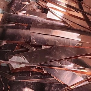 Puede incluir: Una colección de recortes de cobre planos y rectangulares y tiras metálicas delgadas con un acabado reflectante. Las piezas están dispuestas en una pila desordenada, mostrando varias longitudes y anchos con superficies lisas y brillantes.