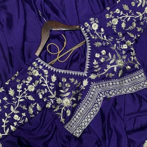 Puede incluir: Un sari de color morado intenso con intrincados bordados florales plateados a lo largo de los bordes y la blusa. La prenda se muestra en una percha de madera, destacando un escote en V y un detallado trabajo de hilo plateado.
