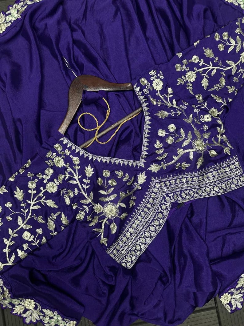 Puede incluir: Un sari de color morado intenso con intrincados bordados florales plateados a lo largo de los bordes y la blusa. La prenda se muestra en una percha de madera, destacando un escote en V y un detallado trabajo de hilo plateado.