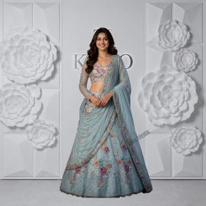 Könnte beinhalten: Ein hellblaues Lehenga-Choli-Set bestehend aus einer langärmeligen Bluse und einem bodenlangen Rock, beide verziert mit aufwendigen Blumenstickereien in Rosa-, Orange- und Grüntönen, ergänzt durch einen passenden transparenten Dupatta über der Schulter.