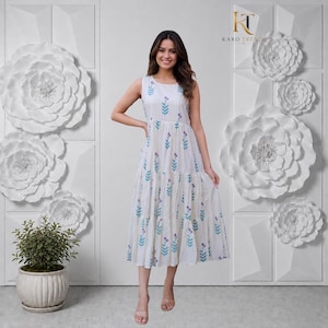 Puede incluir: Un vestido midi blanco sin mangas con falda de niveles y un patrón repetitivo de pequeñas flores azules y moradas. El vestido tiene un cuello redondo y un corpiño ajustado que se transforma en una falda fluida con niveles fruncidos.