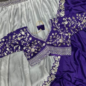 Puede incluir: Un conjunto de dos piezas que incluye una falda de sat&eacute;n plateado con un borde ancho bordado y una blusa de color morado intenso. La blusa tiene cuello en V, mangas tres cuartos y un bordado floral plateado con lentejuelas.