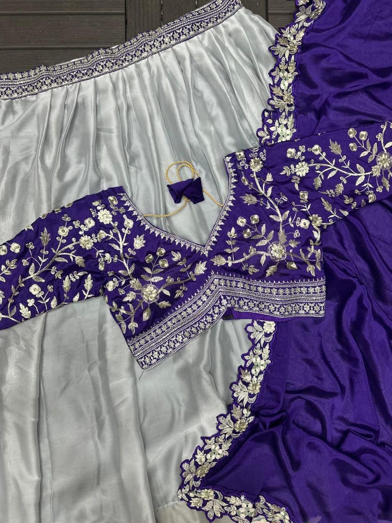 Puede incluir: Un conjunto de dos piezas que incluye una falda de sat&eacute;n plateado con un borde ancho bordado y una blusa de color morado intenso. La blusa tiene cuello en V, mangas tres cuartos y un bordado floral plateado con lentejuelas.
