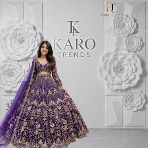 May include: A deep purple lehenga choli featuring intricate gold embroidery throughout the skirt and blouse. The ensemble includes a matching purple sheer dupatta with a gold border. The design showcases detailed patterns and a flared silhouette.