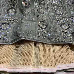 May include: A sage green skirt featuring intricate silver embroidery and circular mirror embellishments arranged in floral patterns. The garment includes a layered beige mesh underskirt with a white hemline, showcasing detailed needlework.
