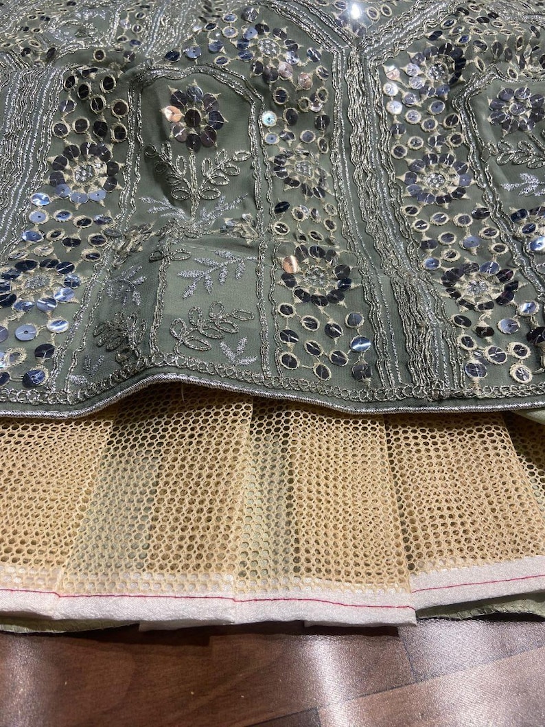 May include: A sage green skirt featuring intricate silver embroidery and circular mirror embellishments arranged in floral patterns. The garment includes a layered beige mesh underskirt with a white hemline, showcasing detailed needlework.