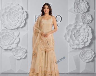 Puede incluir: Un conjunto de kurta y sharara beige sin mangas con intrincados bordados de motivos florales y de pavo real, combinado con una dupatta transparente a juego. La prenda muestra un detallado trabajo de costura en el dobladillo y el corpiño, con el texto KARO TRENDS.