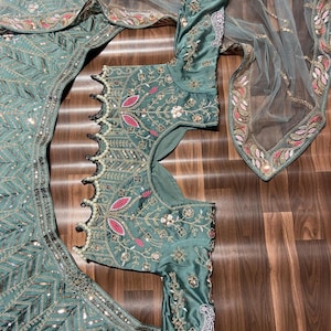 Puede incluir: Un conjunto de lehenga choli verde salvia con intrincados bordados de hilo plateado, lentejuelas y detalles florales rosas. La blusa tiene mangas largas y un dobladillo festoneado adornado con perlas blancas, junto a un dupatta transparente.