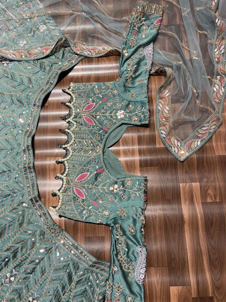 Puede incluir: Un conjunto de lehenga choli verde salvia con intrincados bordados de hilo plateado, lentejuelas y detalles florales rosas. La blusa tiene mangas largas y un dobladillo festoneado adornado con perlas blancas, junto a un dupatta transparente.