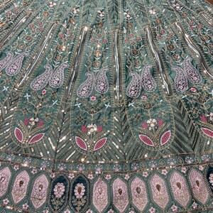 Puede incluir: Una falda lehenga de color verde salvia con intrincados bordados de motivos florales y de cachemira. La tela est&aacute; adornada con hilo met&aacute;lico plateado, lentejuelas y peque&ntilde;os espejos dispuestos en paneles verticales y un borde ancho.