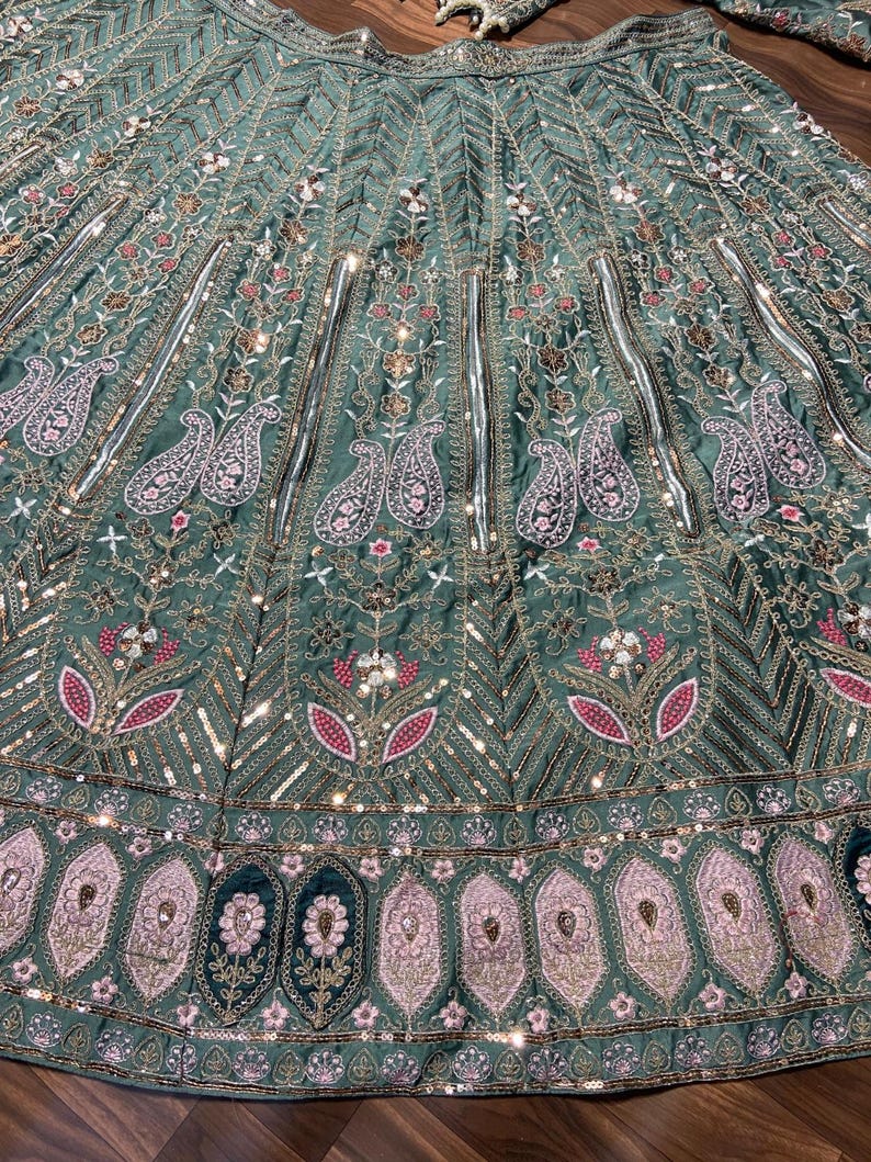 Puede incluir: Una falda lehenga de color verde salvia con intrincados bordados de motivos florales y de cachemira. La tela est&aacute; adornada con hilo met&aacute;lico plateado, lentejuelas y peque&ntilde;os espejos dispuestos en paneles verticales y un borde ancho.