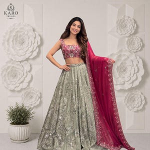 May include: A two-piece lehenga set featuring a sleeveless, fuchsia-pink cropped blouse with intricate floral embroidery and a high-waisted, floor-length silver skirt adorned with extensive mirror work and a matching fuchsia-pink sheer dupatta.