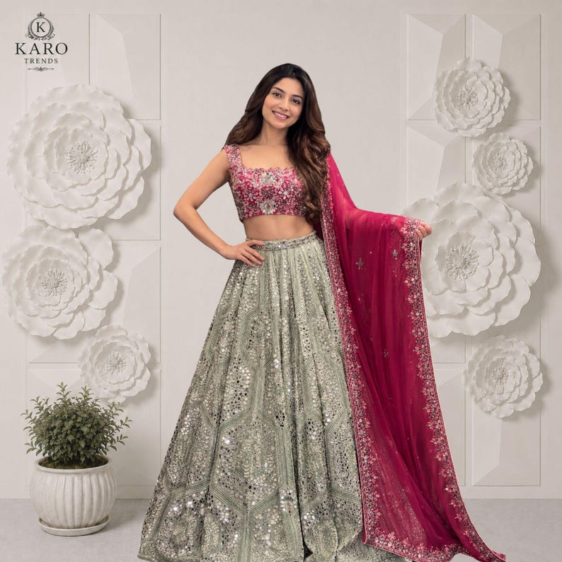 May include: A two-piece lehenga set featuring a sleeveless, fuchsia-pink cropped blouse with intricate floral embroidery and a high-waisted, floor-length silver skirt adorned with extensive mirror work and a matching fuchsia-pink sheer dupatta.