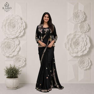 May include: A black saree paired with a matching black jacket featuring floral embroidery on the shoulders and sleeves. The saree includes a decorative border with small square accents and intricate floral embroidery along the hemline.