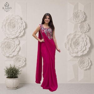 Puede incluir: Un sari de color magenta vibrante con un corpiño sin mangas que presenta un intrincado bordado floral en tonos azul, rosa y verde. La prenda incluye un largo drapeado fluido que cae desde el hombro hasta el suelo, creando una silueta limpia.